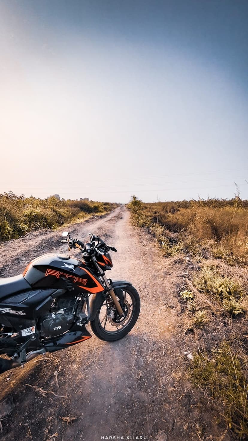 Black and orange motorcycle parked on a dirt trail through dry grass under a glowing orange sunset sky.