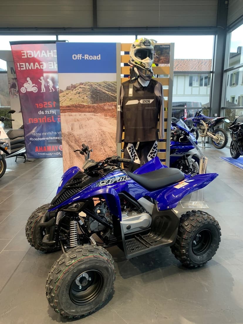 Blue Yamaha YFM90R off-road quad bike showcased in a bright dealership with knobby tires and motocross gear.