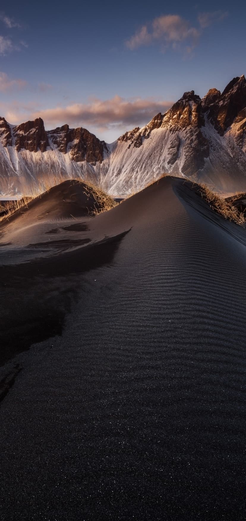 Dark sand dunes with wind ripples sit before glowing snow-capped mountains under a soft blue and orange dusk sky.
