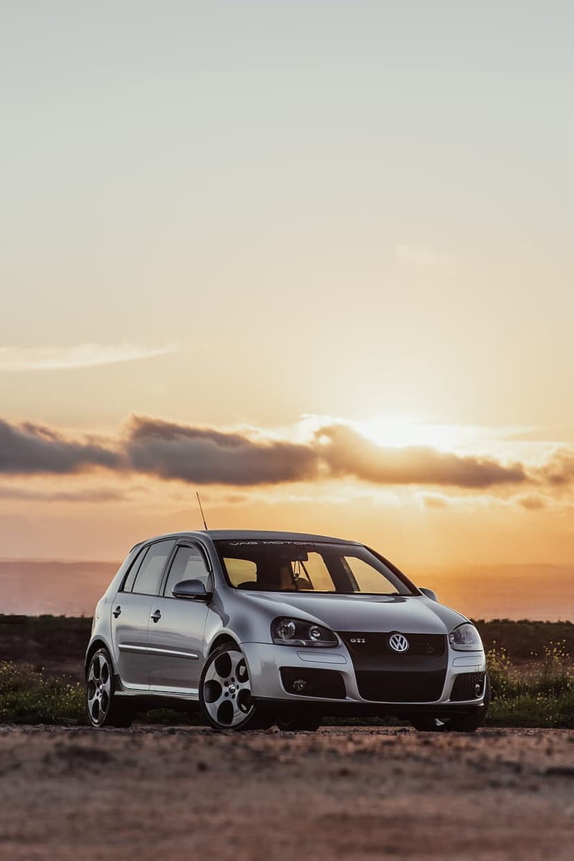 Silver Volkswagen Golf GTI Mk5 parked on a dirt path under a dramatic orange sunset with dark clouds.