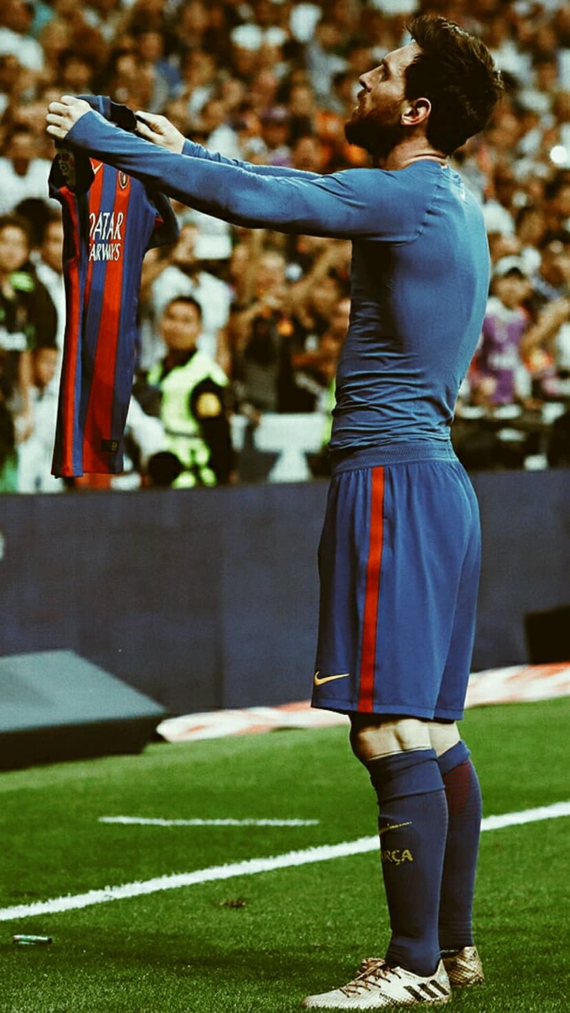 Lionel Messi stands on a football pitch holding up a blue and red striped jersey toward a cheering crowd.