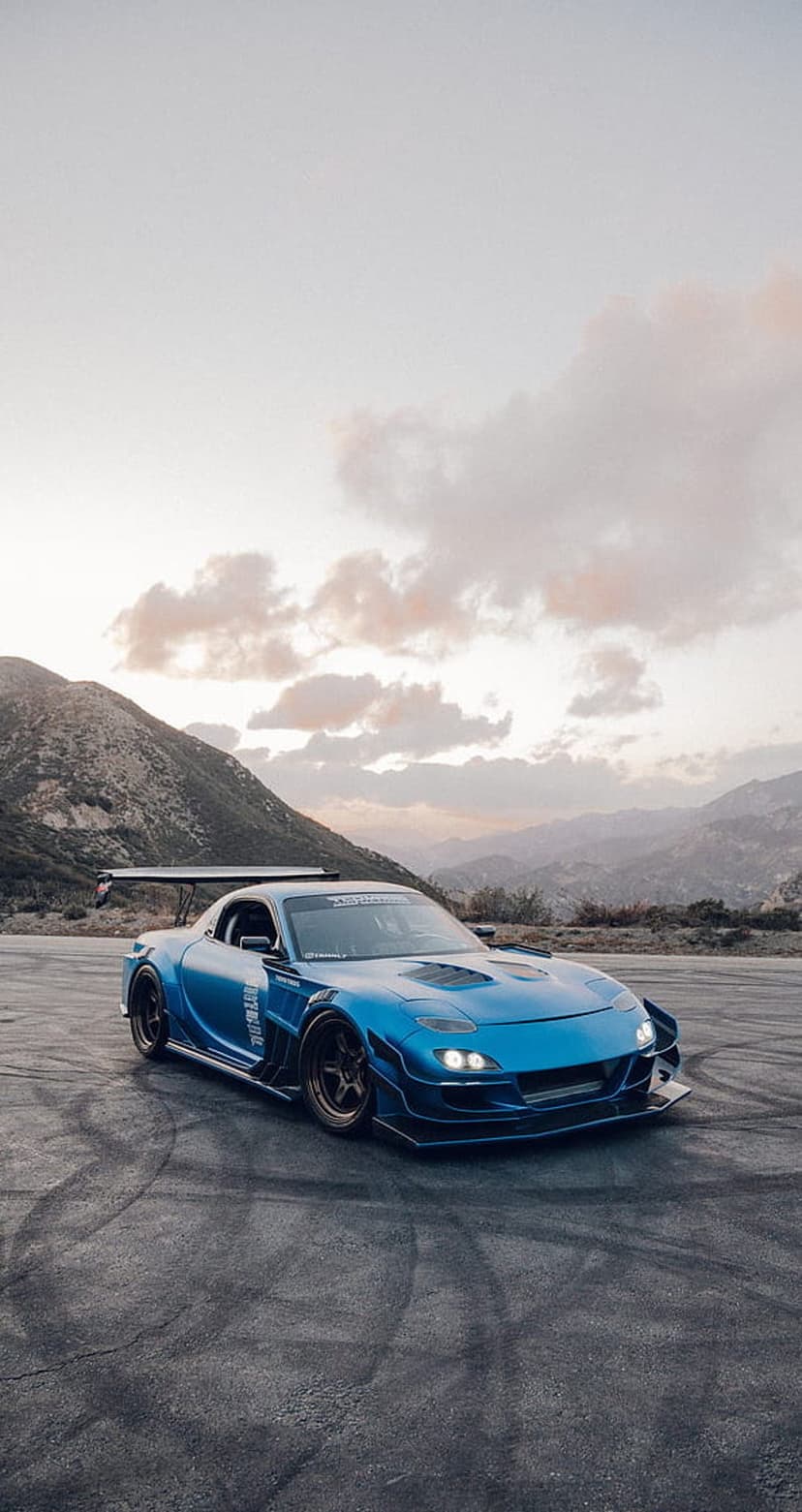 Blue Mazda RX-7 with a widebody kit drifting on a mountain road during a vibrant orange and purple sunset.