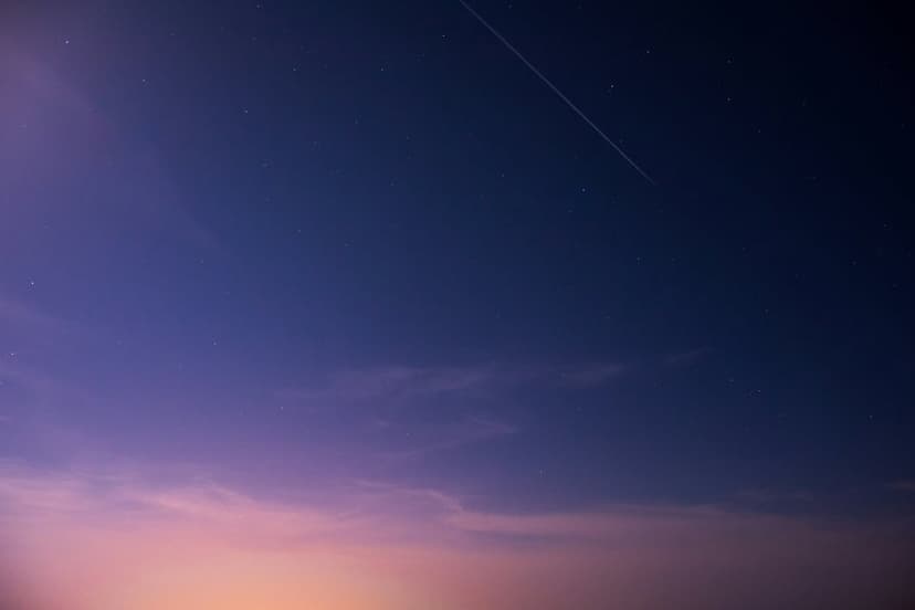 Bright meteor trail bisects a gradient dusk sky of purple and blue above a layer of hazy evening clouds.