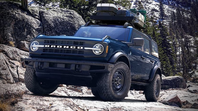 Blue Ford Bronco climbing a rocky slope with a roof rack of gear against a snowy pine forest background.