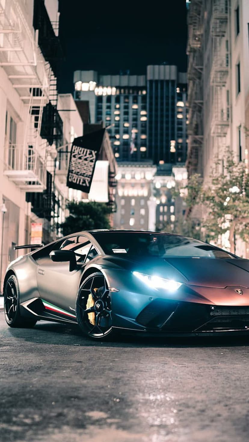 Lamborghini supercar with glowing headlights parked on wet city pavement under glowing urban building lights.