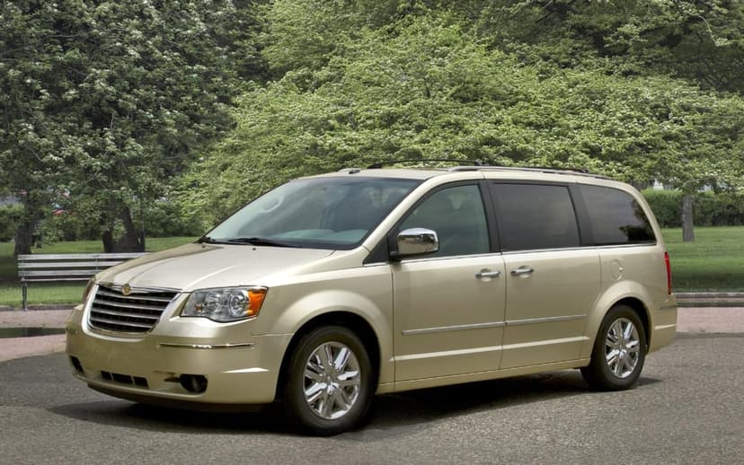 Gold Chrysler Town and Country minivan parked on asphalt against a backdrop of dense green trees in daylight.