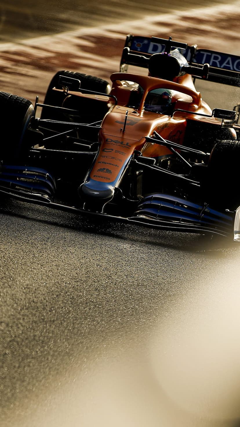 Orange McLaren F1 car front wing and tires on a textured racetrack asphalt surface under bright sunlight.