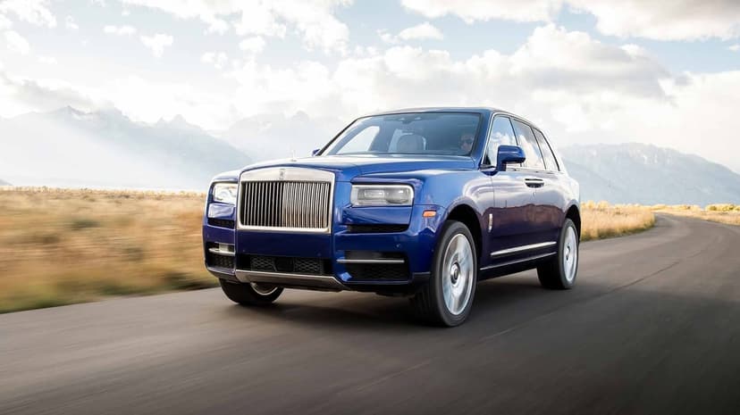 Blue Rolls-Royce Cullinan driving on an open road with misty mountains and a cloudy sky in the background.