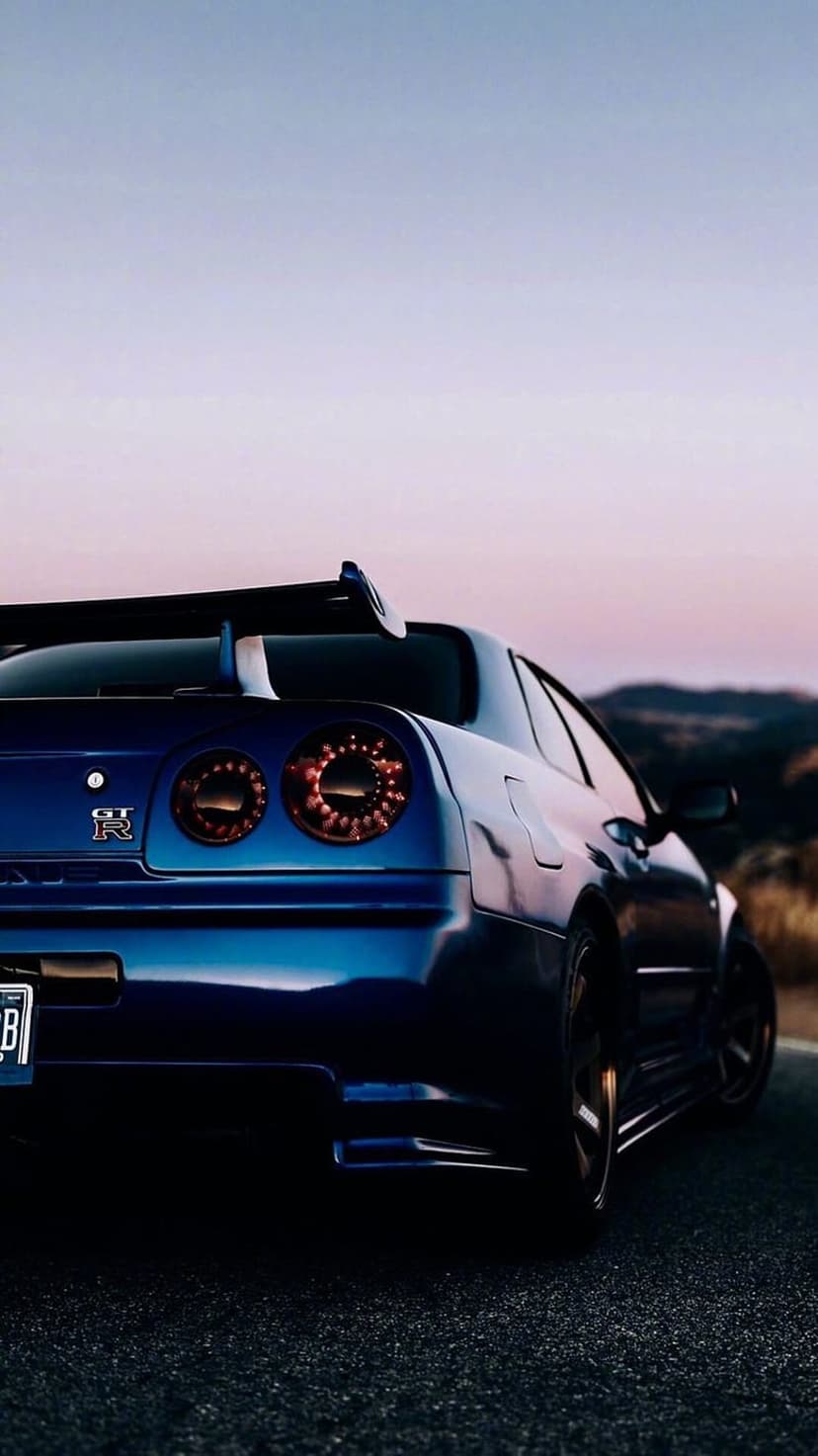 Nissan GT-R R34 circular red tail lights glowing on a blue car body during a colorful mountain sunset landscape.