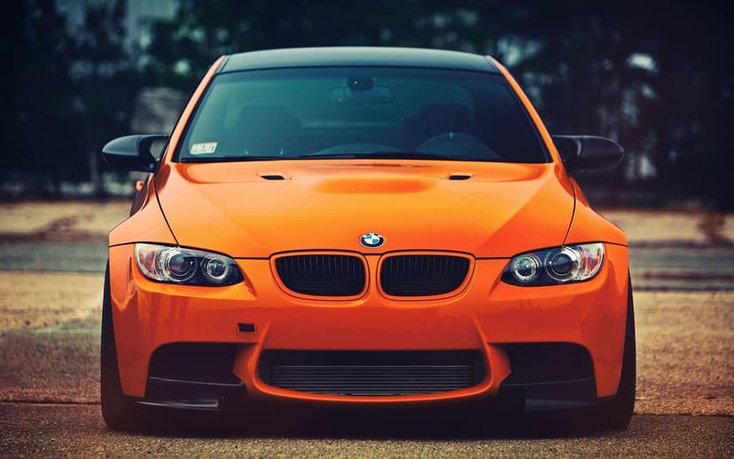 Front view of an orange BMW M3 E92 with black grilles and carbon fiber accents on a blurred road background.