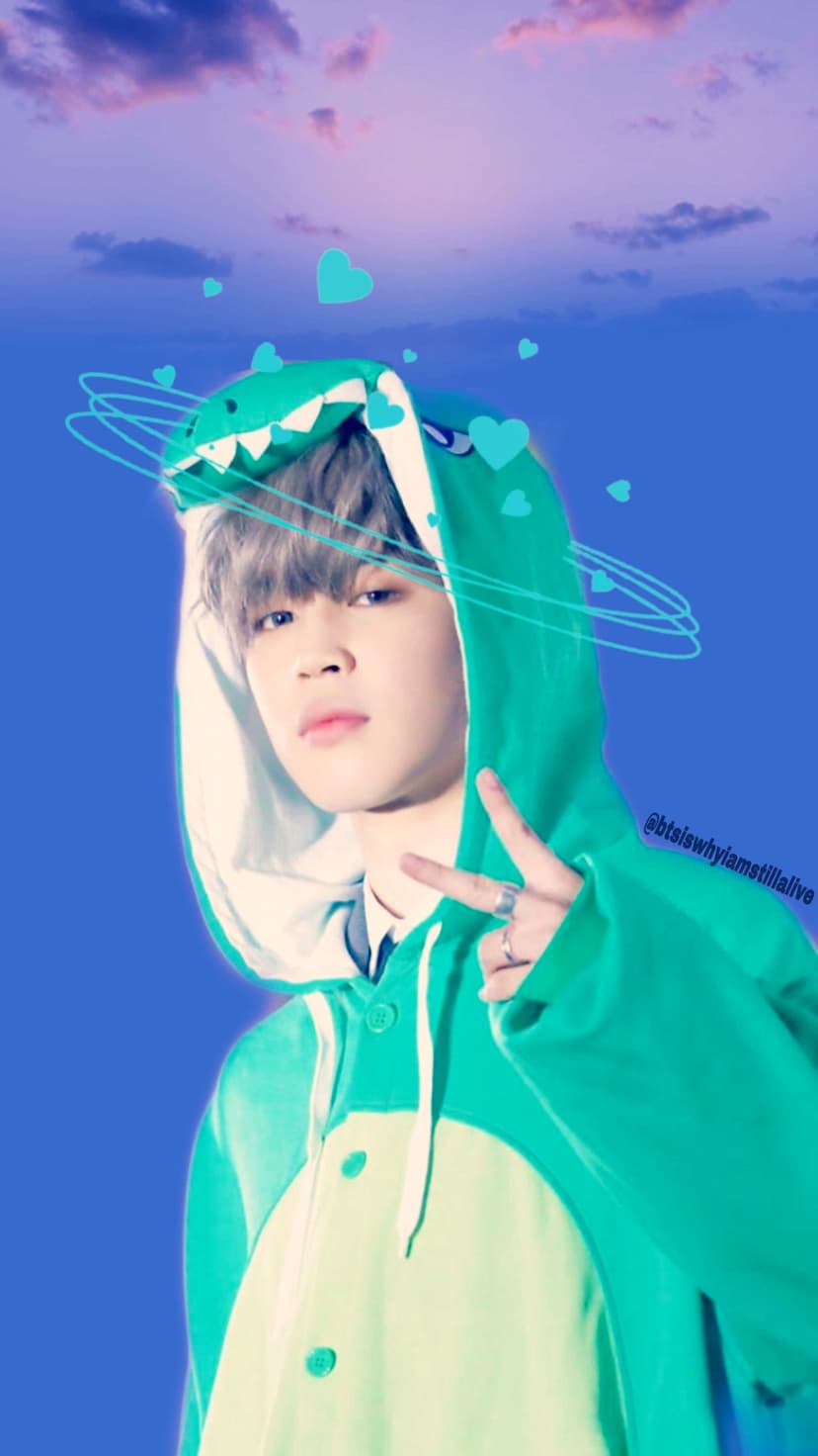 Park Jimin wears a green dinosaur kigurumi under a purple sky with floating teal hearts and light blue swirls.