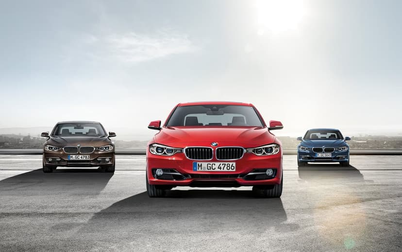 Three BMW 3 Series cars in red, brown, and blue parked on asphalt under a bright sunny sky with lens flare.