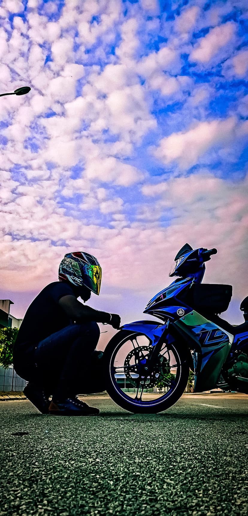 Motorcyclist in a bright helmet crouches beside a blue sportbike under a vast, dramatic white and blue cloudy sky.