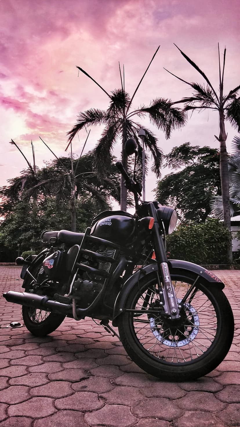 Black Royal Enfield motorcycle on a cobblestone path against a pink sunset sky with palm tree silhouettes.