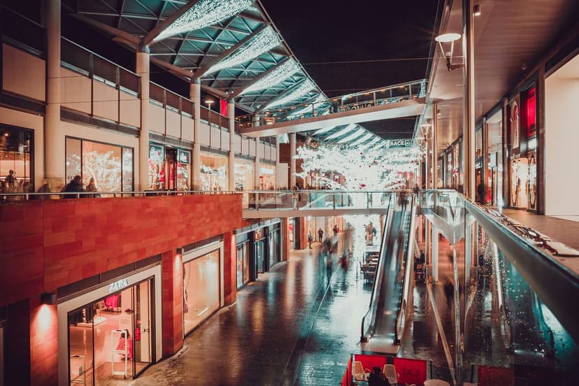 Illuminated escalators traverse a multi-story shopping mall interior with glowing storefronts and glass ceilings.