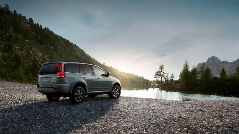 Silver SUV parked on a pebbly shore next to a calm lake with mountains and pine forests under a golden sunset.