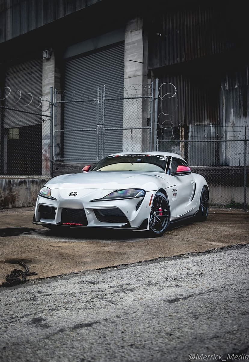 White Toyota Supra sports car with red accents parked near a chain-link fence and weathered industrial building.