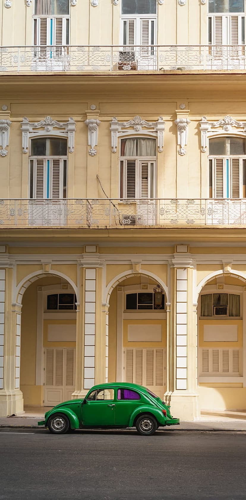 Green Volkswagen Beetle parked on a historic Havana street in front of a pale yellow building with arched windows.