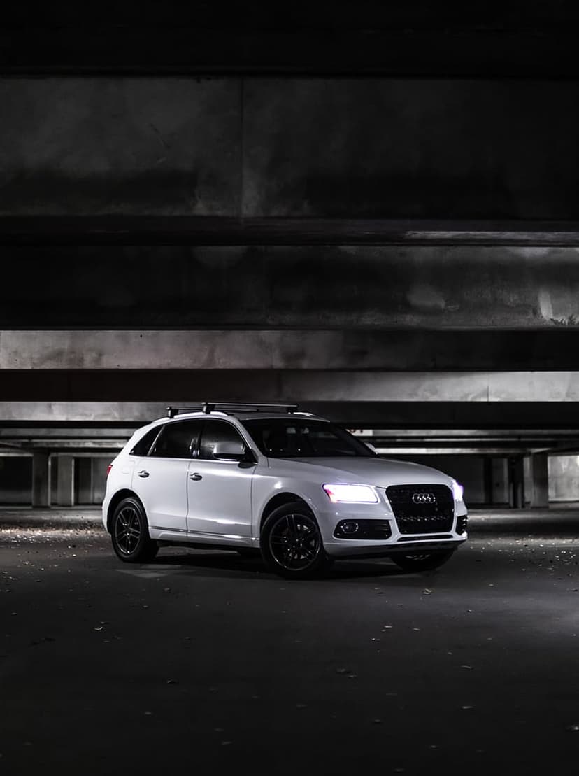 White Audi Q5 SUV with black rims and roof rack parked in a dark concrete garage with its headlights illuminated.