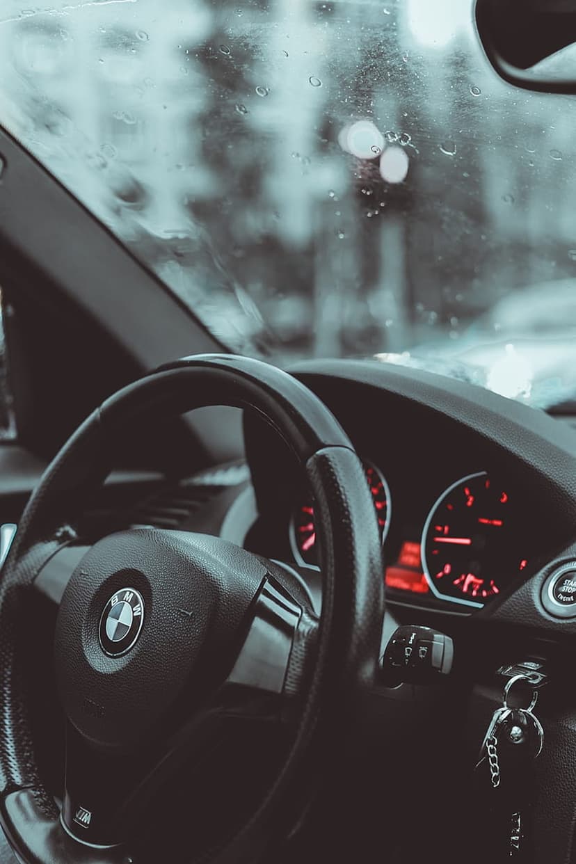 BMW steering wheel and digital dashboard glow red behind a glass windshield covered in heavy raindrops.