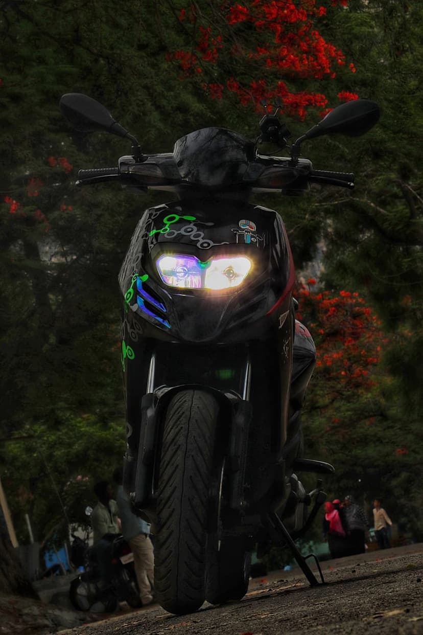 Black scooter with glowing headlights on a dirt road surrounded by lush green trees and red flowers at dusk.
