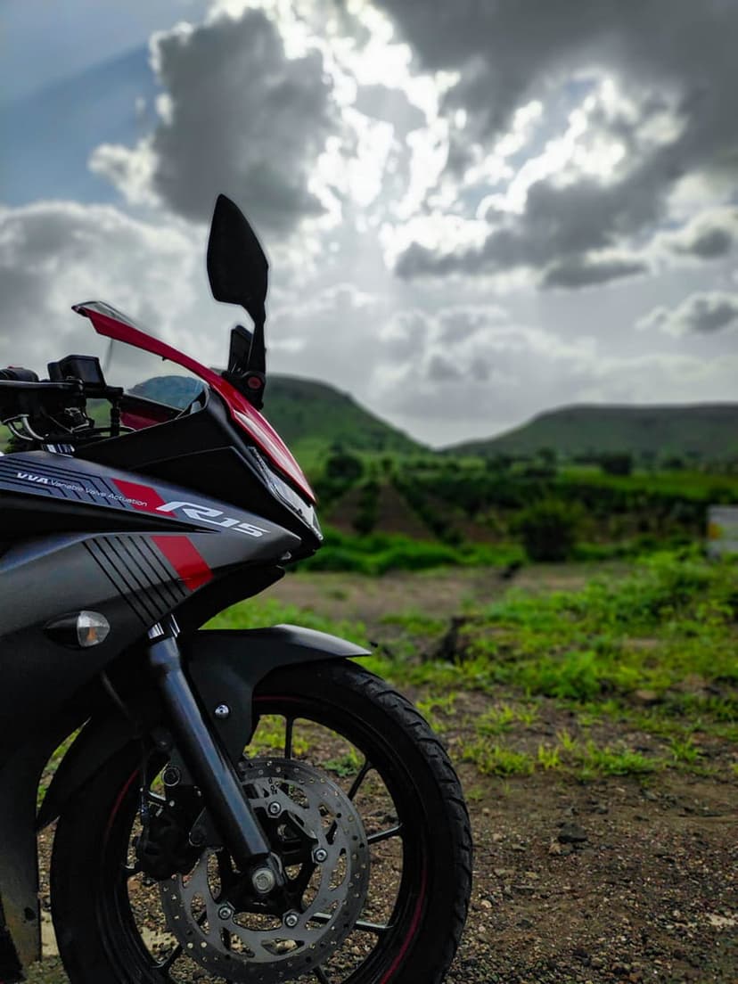 Yamaha R15 motorcycle with grey and red paint parked on a dirt road with green hills and stormy clouds behind it.