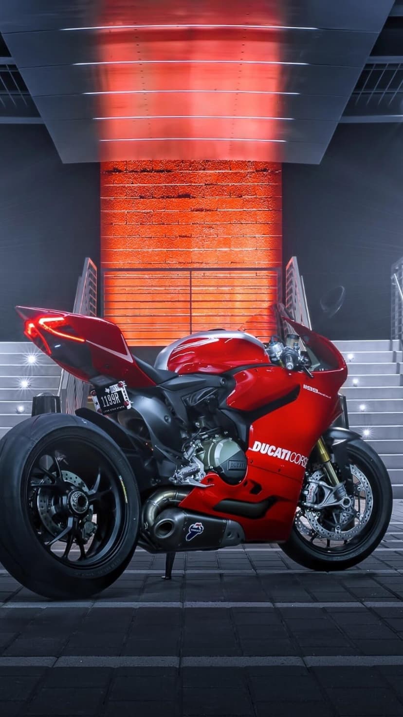 Red Ducati Corse sportbike parked on dark tiles under glowing crimson overhead lights in an urban setting.