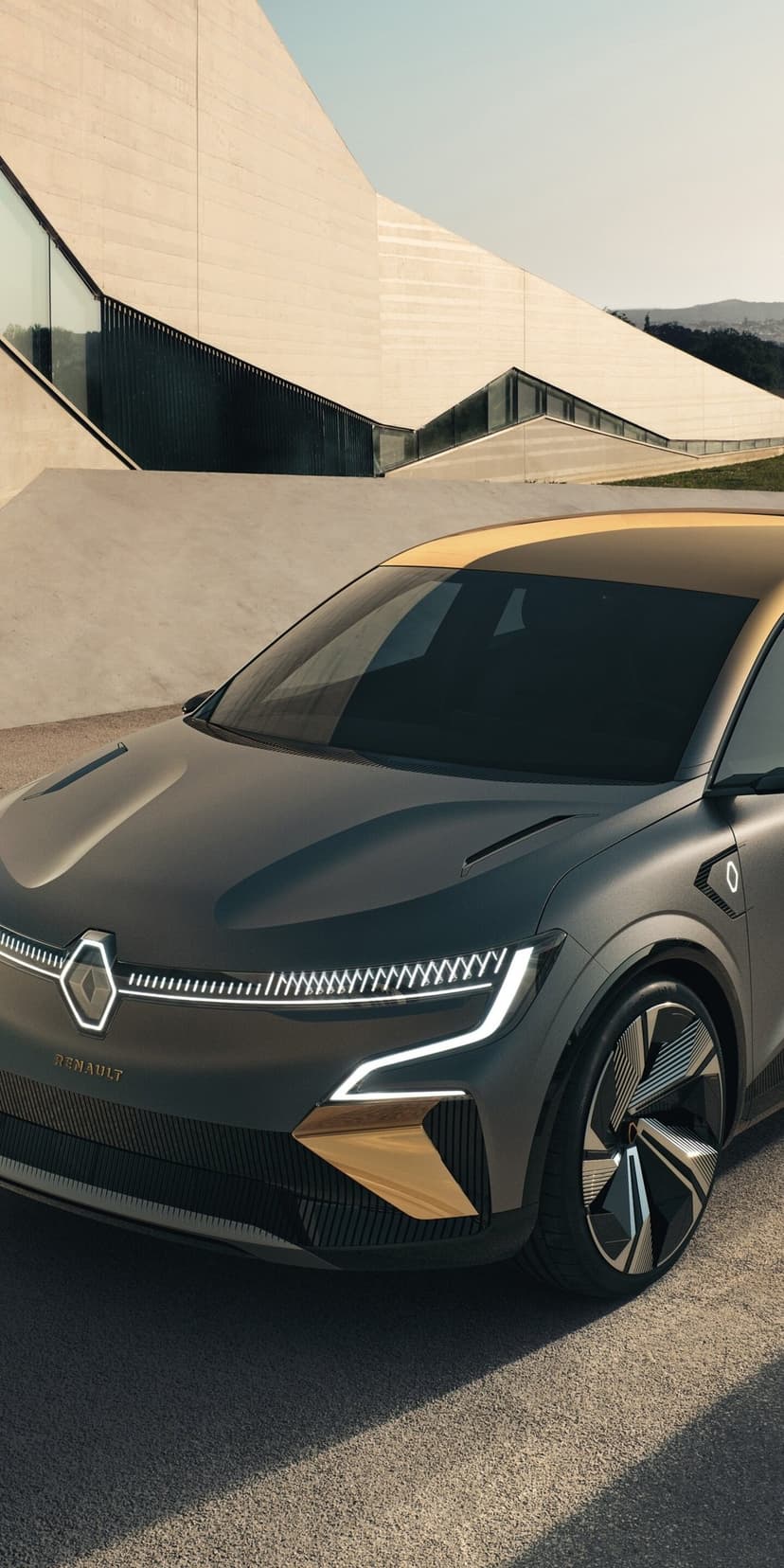 Front view of a matte grey electric car with gold trim and glowing LED lights near modern concrete buildings.