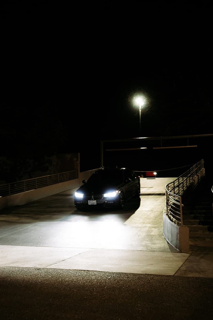 Black BMW sedan drives through a dark concrete parking garage with bright headlights and an overhead streetlamp.