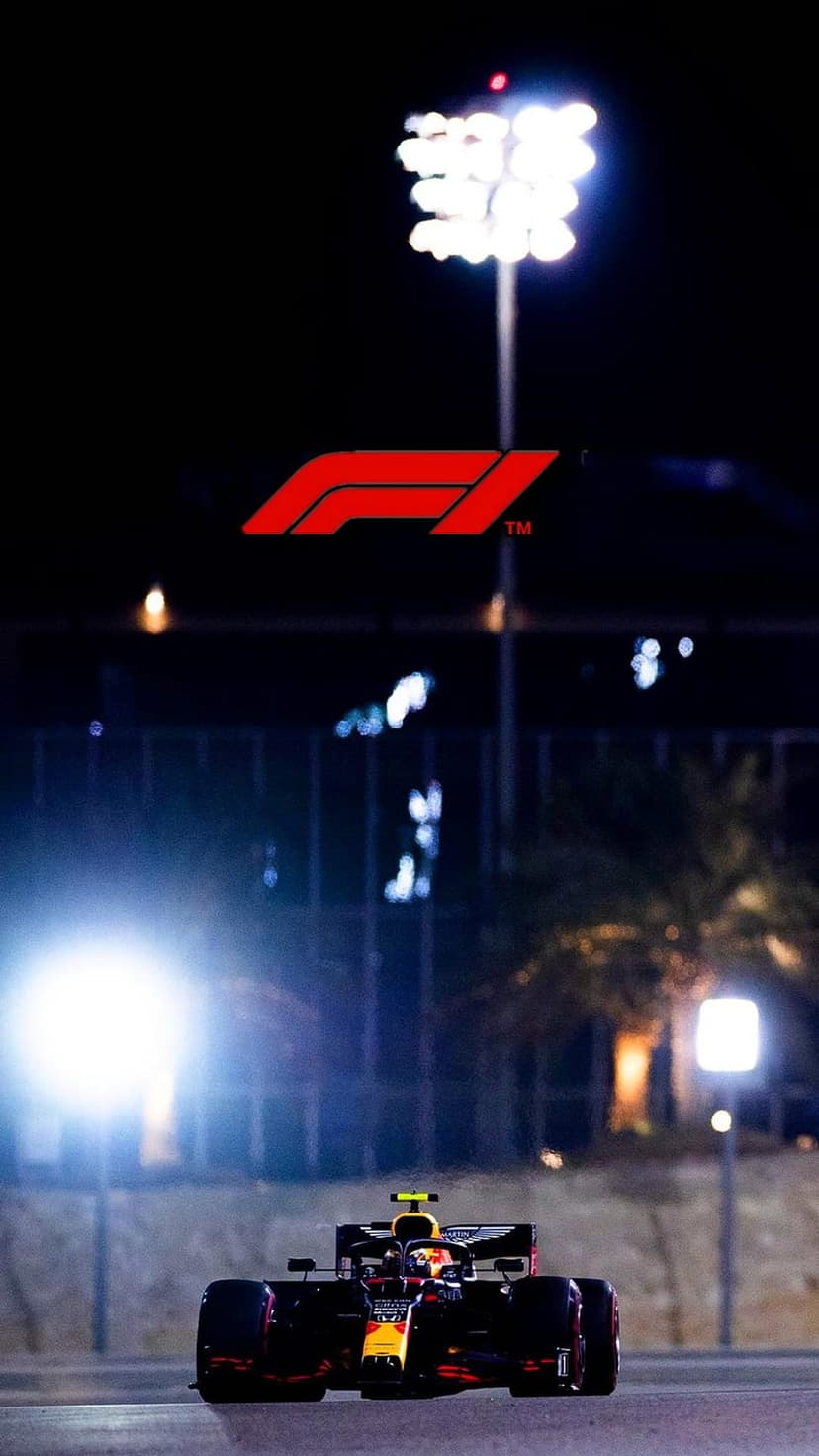 Formula 1 race car in red and yellow speeds past floodlights on a night track with a visible logo above.