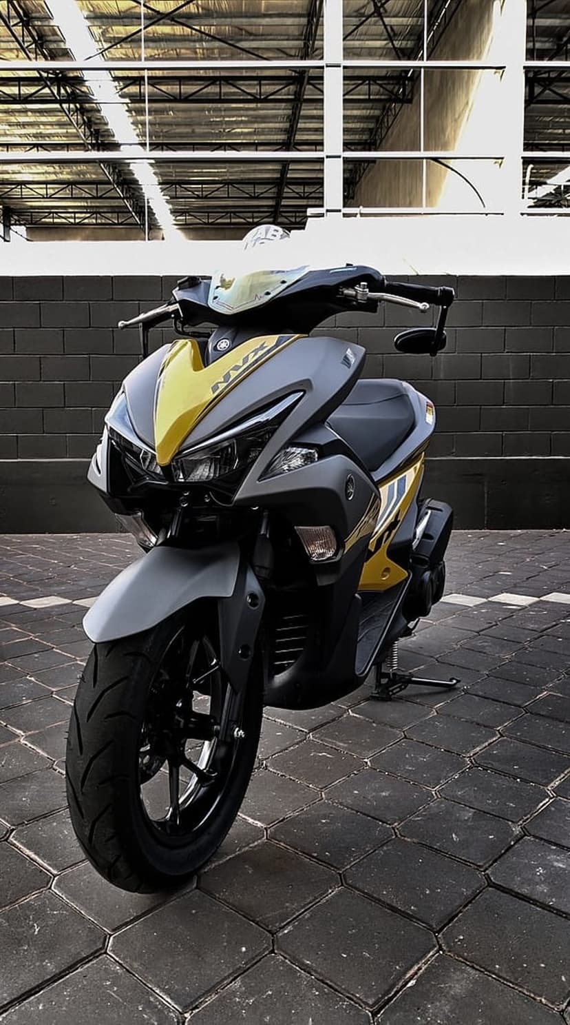 Yellow Yamaha NVX scooter parked by a brick wall with gray accents and sharp aerodynamic fairings on paved ground.