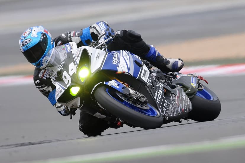 Professional racer on a blue Yamaha R1 leans into a sharp turn at high speed on an asphalt racing circuit.