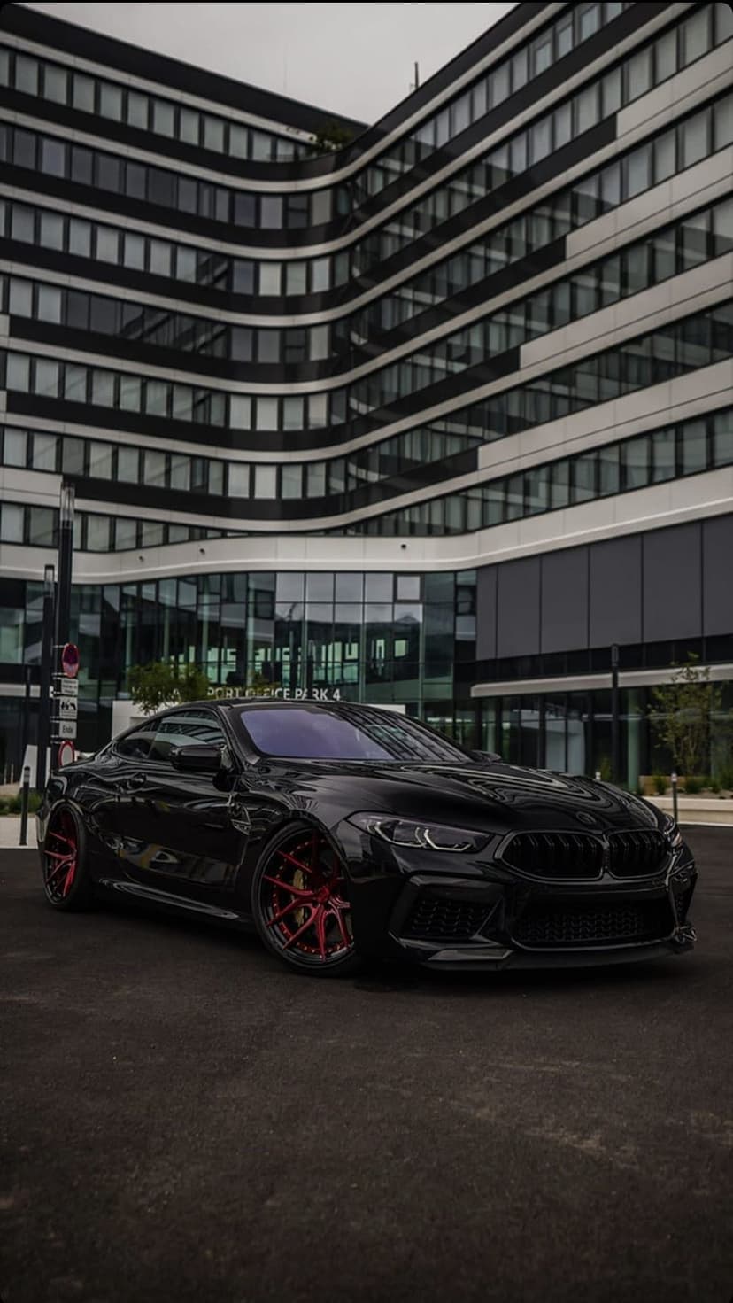 Black BMW M8 Competition with red aftermarket wheels parked in front of a modern glass building in an urban setting.