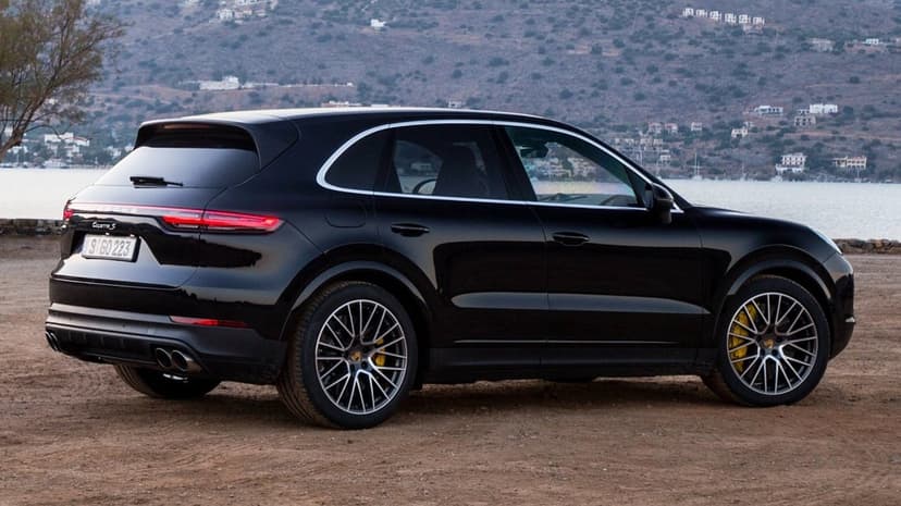 Black Porsche Cayenne S SUV on dirt ground with yellow brake calipers and a coastal hillside background.