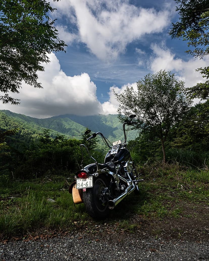 Black cruiser motorcycle parked on a grassy mountain road overlooking green hills and dramatic white clouds.