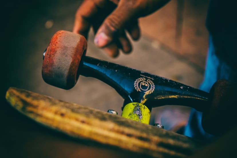 Skateboard truck with a gold smiley logo and red wheel sits on a wooden deck as a hand touches the hardware.