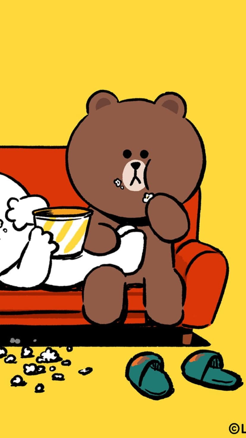 Brown bear and white character share popcorn on a bright orange sofa with teal slippers on the floor.