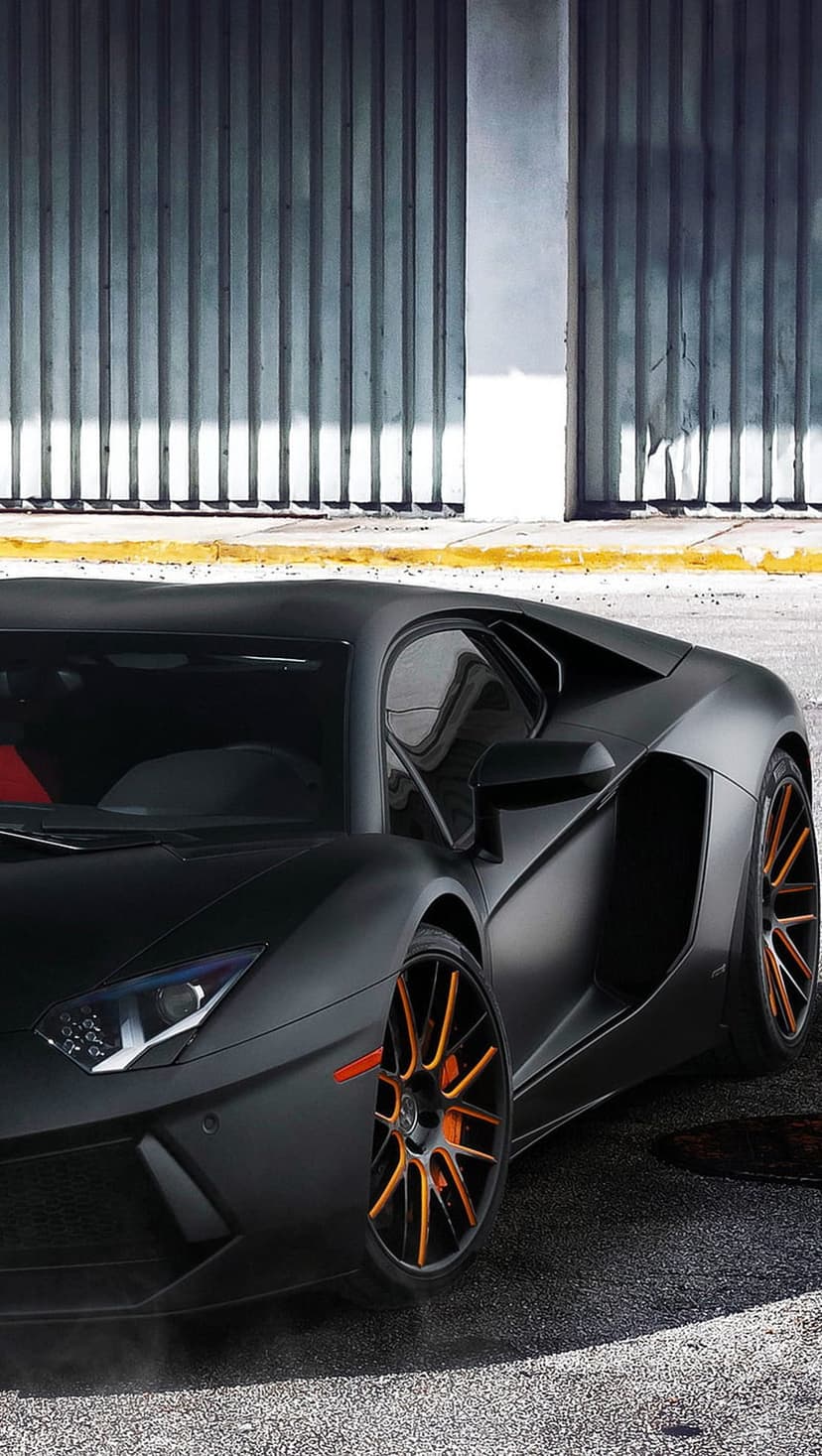 Lamborghini Aventador in matte black with orange wheels parked in front of a corrugated metal garage door.