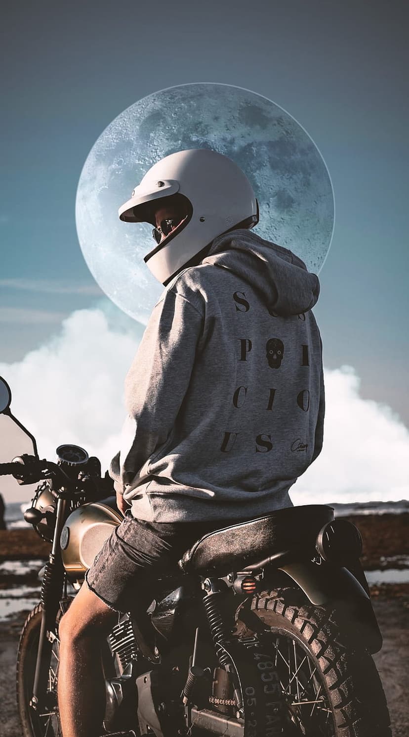 Motorcyclist in a white helmet and skull hoodie sits on a bike facing a giant moon through cloudy night skies.
