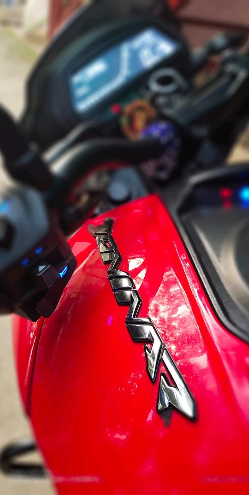 Red motorcycle RS logo with chrome finish on a crimson fairing, featuring a blurred dashboard in the background.