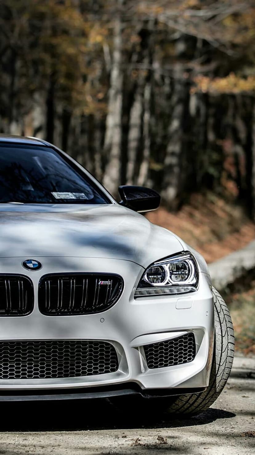 White BMW M6 front grille and headlight on a dirt road with blurred orange autumn trees in the background.
