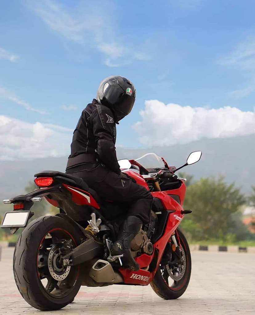 Motorcyclist in black gear sits on a red sportbike against a backdrop of blue sky, white clouds, and green hills.