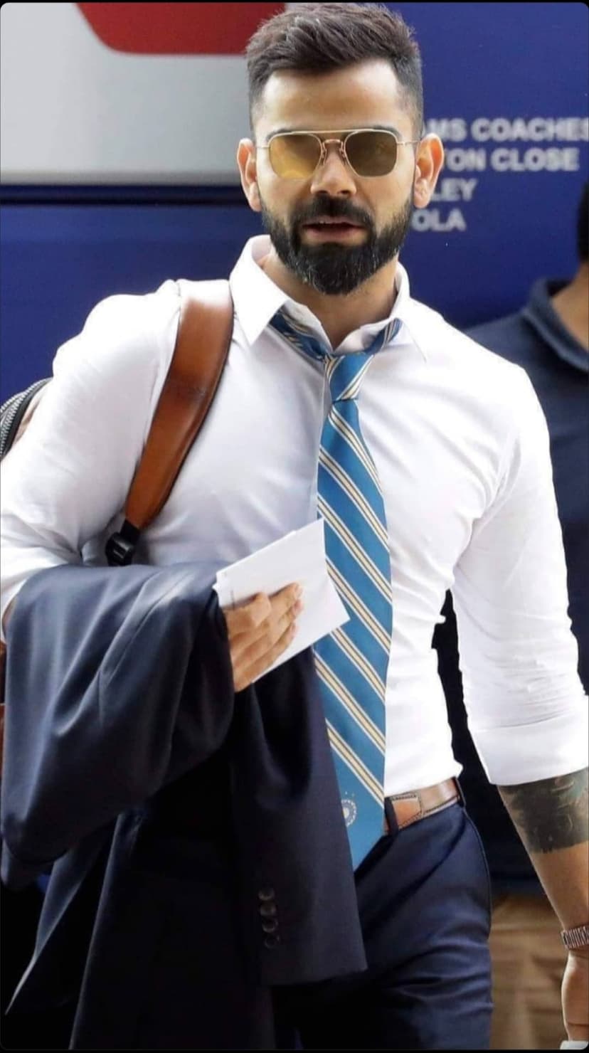 Virat Kohli in a white shirt and striped tie wears aviator sunglasses and carries a dark blazer and brown bag.