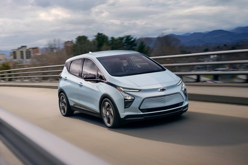 Light blue electric Chevrolet Bolt EV driving on a bridge with mountains and an overcast sky in the background.