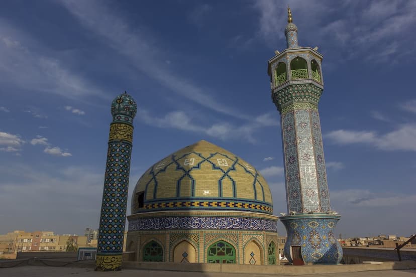 Mosaic mosque dome and minaret with geometric tilework against a bright blue sky with scattered clouds.