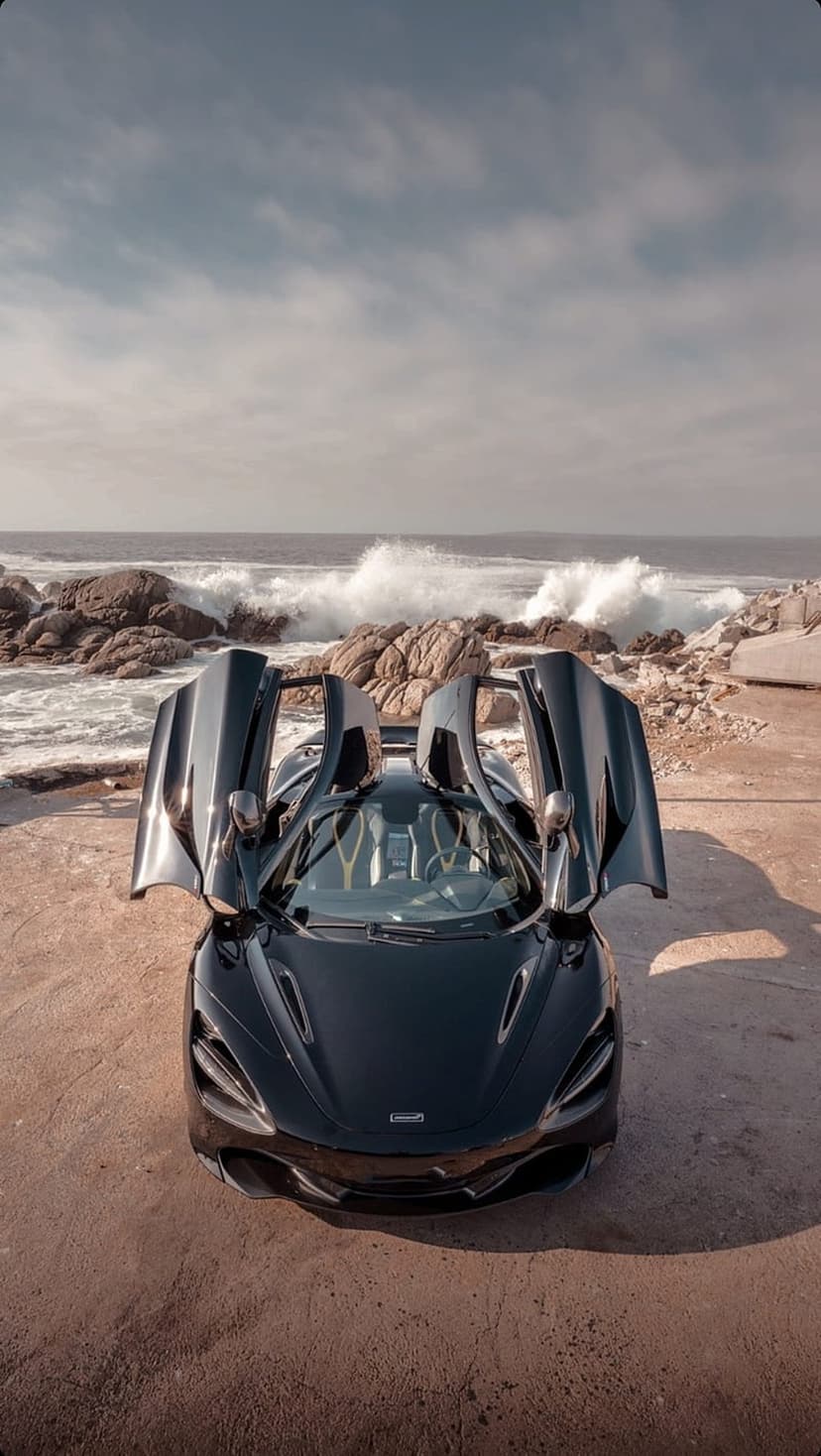 Black McLaren supercar with open doors parked on a sandy beach with crashing ocean waves and coastal rocks.