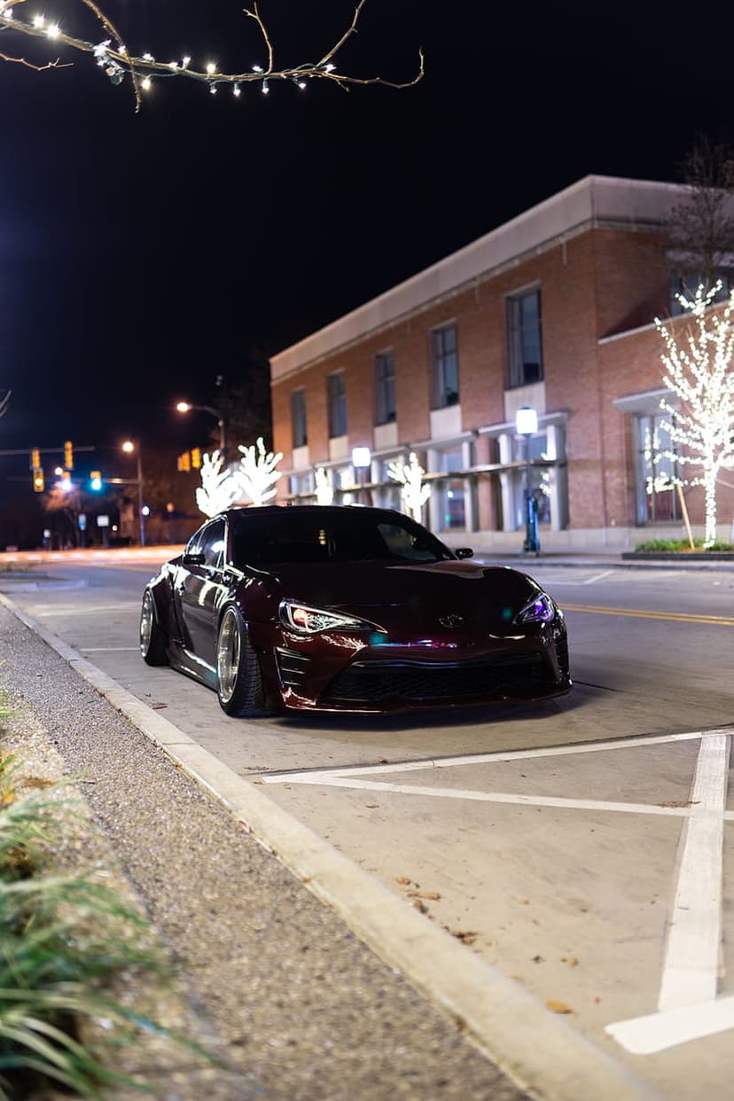 Dark red sports car with wide fenders parked on a night street illuminated by festive overhead lights.