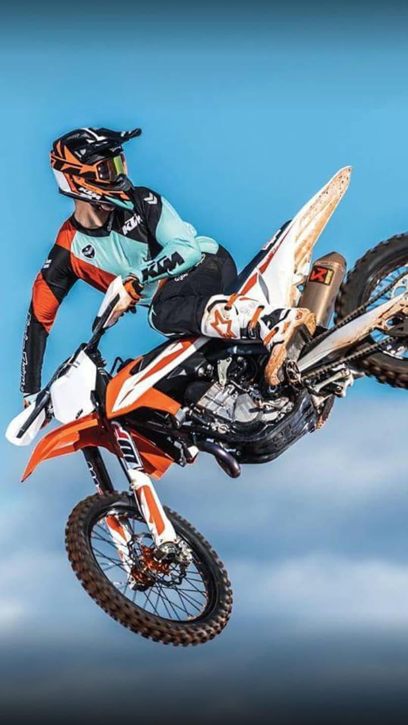 Motocross athlete on an orange dirt bike performs an aerial stunt against a vivid blue sky with thin clouds.