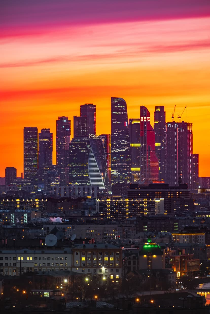 Modern glass skyscrapers stand tall against a fiery orange and purple sunset sky during the golden hour.