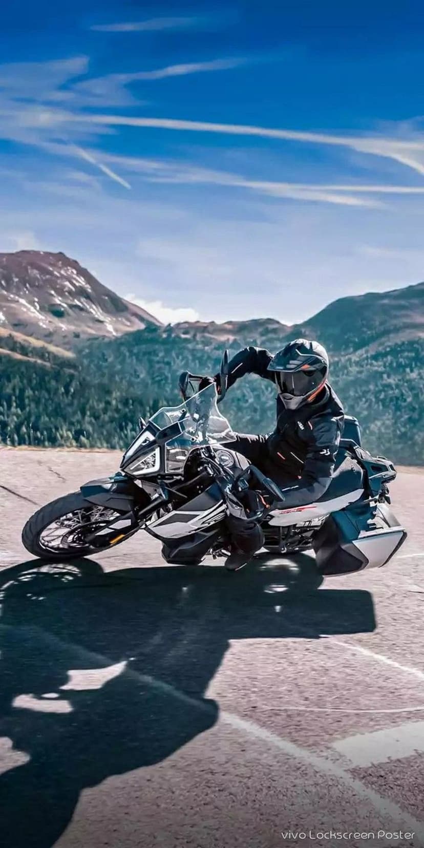 Motorcyclist leaning into a paved road turn with snow-capped alpine mountains and a blue sky in the background.